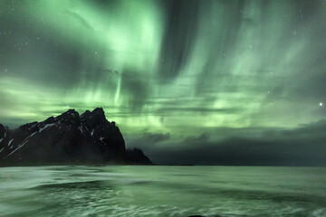 View of Aurora Borealis dances above the tranquil sea and silhouetted mountains, casting an ethereal green glow over the serene landscape, Hofn, Iceland.