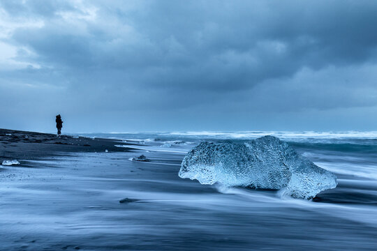 View of a glistening ice diamond rests on the black sand beach as waves surge creating a blurred effect, under a dramatic sky, Iceland.