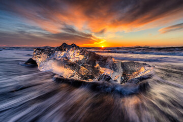 View of a luminous iceberg fragment rests on the black sand beach as the tide rushes around, a vibrant sunset paints the sky, Iceland.