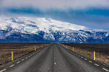 View of an endless asphalt road stretches towards a majestic snow-capped mountain range under a cloudy sky, inviting exploration and adventure, Iceland.