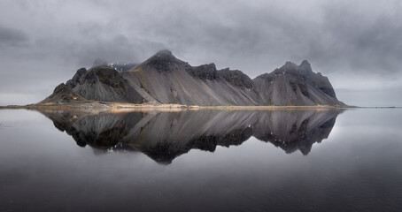 View of stark, jagged mountains mirroring in the still, dark water under a brooding sky, an ethereal landscape, Hofn, Iceland.