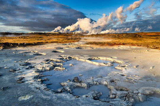 View of geothermal area with bubbling mud pools, steaming vents, and contrasting colors under a dynamic sky, Hverir, Iceland.