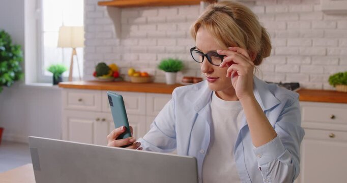 Young woman wearing eyeglasses suffering from presbyopia sitting in kitchen at home trying to read message on mobile phone. Farsightedness eye disease and vision problems. 4k video.
