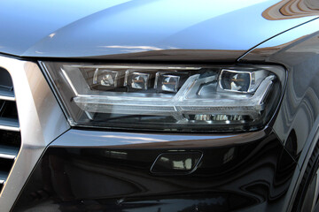 Close-up of a modern black cars right LED headlight and chrome grille with a glossy body surface reflecting urban surroundings, showcasing sleek lines and high-end exterior detailing. © Best Auto Photo