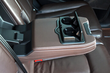 Luxury rear car seat with open brown leather armrest featuring a dual black plastic cupholder....