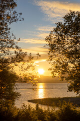 Beautiful sunset over the mountains above the lake. Reflection of sun rays in the water in a sea bay with trees
