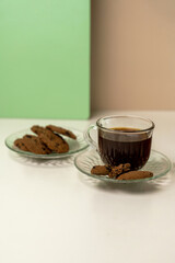A glass cup of black coffee with chocolate chip cookies on a white table. Minimalist still life with a pastel green and beige background.