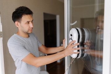 Young handsome man installing robotic vacuum cleaner on window, preparing to activate device for...