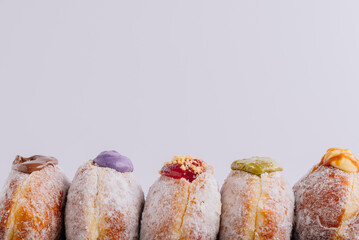 A minimalistic front-facing shot of five assorted sugar-coated doughnuts, each filled with different colorful creams and toppings including chocolate, ube, strawberry crumble, matcha, and mango custar