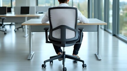 Young Asian businessman sitting alone at desk in modern bright office space with large windows, viewed from behind in ergonomic mesh chair.