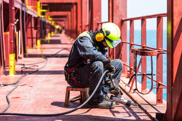 A crew member working on the deck of a merchant ship, using a pneumatic tool to remove rust.