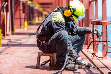 A crew member working on the deck of a merchant ship, using a pneumatic tool to remove rust.