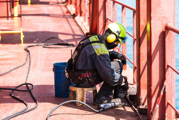 A crew member working on the deck of a merchant ship, using a pneumatic tool to remove rust.