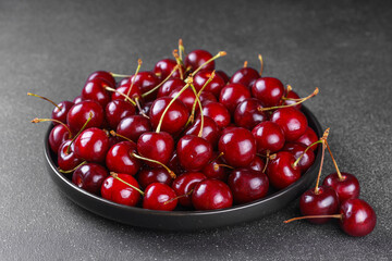 Close up of black plate filled with ripe red sweet cherry placed on dark gray textured background....