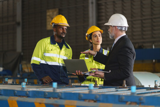 A man and a woman worker are standing in a factory, looking at a tablet. The engineer talks with the woman's workers. They wear a yellow and white safety helmet for safety.