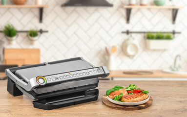 A modern electric grill sits on a wooden countertop beside a plate of freshly grilled chicken breasts with herbs and vegetables in a cozy kitchen