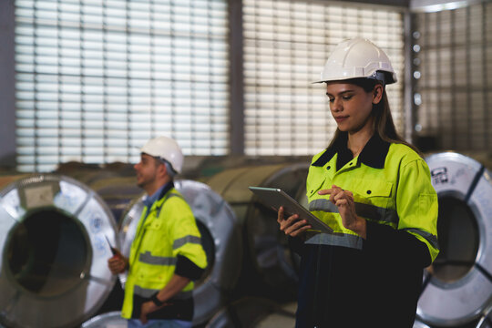 A woman worker wearing a yellow and blue safety vest is holding a tablet in a factory