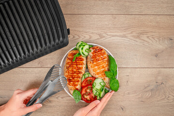 A top-down view shows hands preparing grilled chicken with cucumbers, tomatoes, and basil next to...
