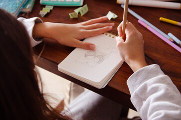 A young girl drawing on a notebook with colorful pencils and some other school supplies on a wooden table.