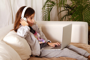 A young girl sitting on a sofa with white headphones and a laptop.