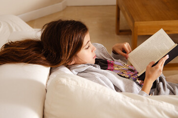 A young girl lying in a sofa reading a book at home. 