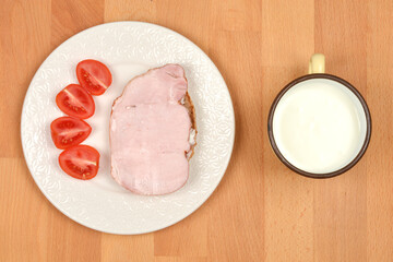 A simple meal is arranged on a light wooden surface, with a slice of ham and four tomato halves on...