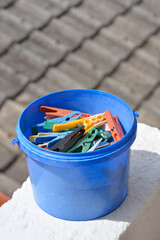 A blue plastic bucket filled with various colorful clothes pins sits outdoors with a blurred tiled...