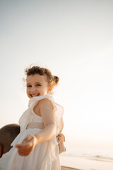 A joyous child, in a white dress, turns and smiles over her shoulder towards the camera on a bright, sunny beach