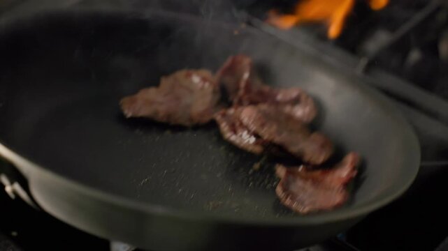 Flame sears raw beef slices in pan on professional gas stove
