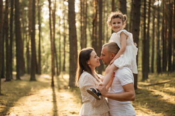 Fototapeta premium A joyous child on father's shoulders, her mother is nearby looking up, all smiling in a sun-drenched pine forest