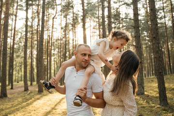 
A joyous child on father's shoulders, her mother is nearby looking up, all smiling in a sun-drenched pine forest