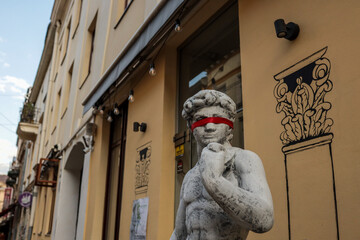 Classical-style sculpture of David with a red blindfold in front of a building with wall art and...