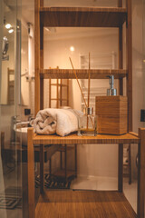 Close-up of a wooden bathroom shelf with a rolled towel, bamboo soap dispenser, and reed diffuser....