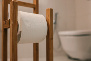 Close-up of a wooden toilet paper holder with white toilet paper roll, modern bathroom in soft focus. White ceramic toilet and bidet sprayer visible in background