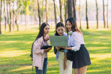 Group of Asian college students standing on green grass and discussing with laptop and books. Concept of teamwork, education, and outdoor learning in a natural environment.