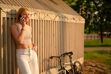 woman enjoying smartphone call outdoors next to bicycle in sunny setting. casual summer attire and...