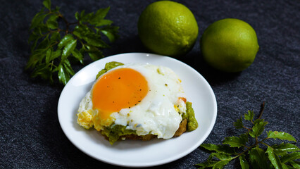 Healthy nutritious breakfast with fried egg sunny side up on toasted bread with mashed avocado and limes on dark background, decorated with green leaves