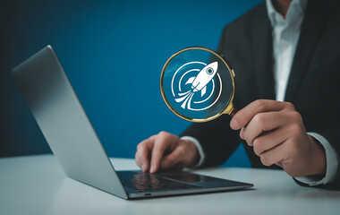 A businessman uses a magnifying glass to examine a rocket launch icon next to a laptop, representing startup research and business opportunities