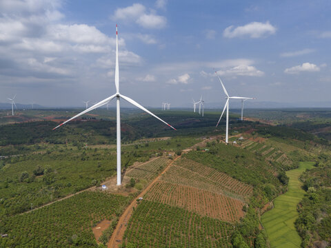 Aerial view of wind turbines standing tall against a backdrop of lush green vegetation and red soil, creating a striking contrast under a partly cloudy sky, Pleiku, Gia Lai, Vietnam.