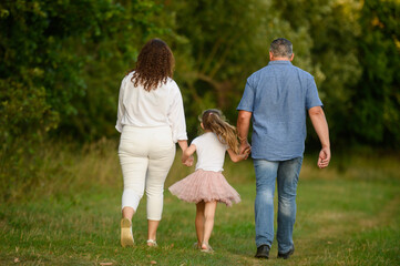 Mom, dad and girl walking holding hands in park and talking, view from back. Girl is jumping...