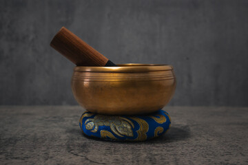 Golden Tibetan singing bowl standing on a small blue cushion promoting relaxation and meditation against a gray backdrop