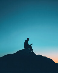 Silhouette of a person sitting on a rock, reading against a clear twilight sky with a gradient from blue to orange.