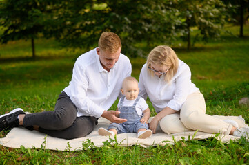 Fototapeta premium Family spending time in park sitting on grass. Mom, dad and baby spending time, parents tickling baby, smiling happily. Family weekend