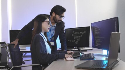 An Asian female developer and an Indian male colleague collaborating on a project. A senior programmer mentors a junior developer in a modern tech office at night. - Powered by Adobe
