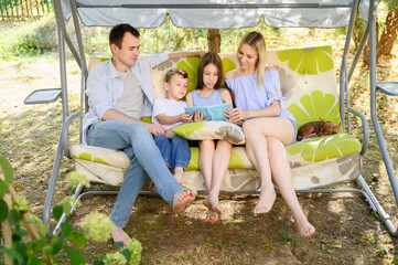 Girl, boy, parents sitting in shade swing swaying reading, discussing what read in book, enjoying...