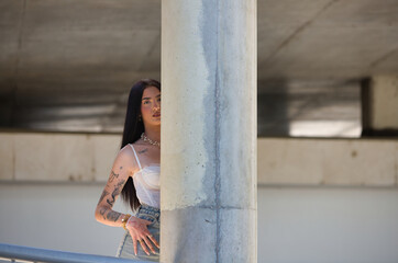 A young, attractive Latin woman with tattoos on her arms and dressed in casual clothes hides behind...