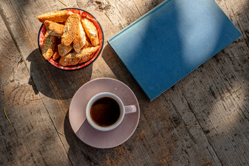 Flat lay of coffee, biscotti and a book on a rustic wooden table with sunlight