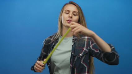 Woman measuring with tape against isolated blue wall showing precision and focus in casual attire, reflecting practicality and concentration in a simple studio setting.