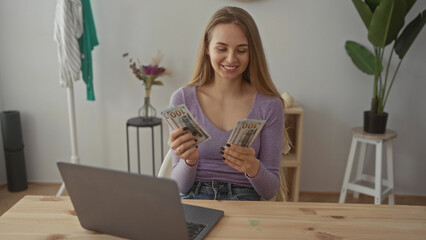 Woman smiling while counting us dollars in bright living room with laptop on wooden table...