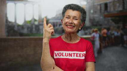 Woman with grey hair wearing volunteer shirt smiling in an urban street setting, pointing upward with her finger, representing senior volunteerism in an old town environment outdoors.